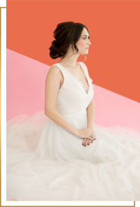 Woman in Bridal Gown Poses on Pink and Choral Background with Low Coiffed Updo with Soft Wavy Tendrils